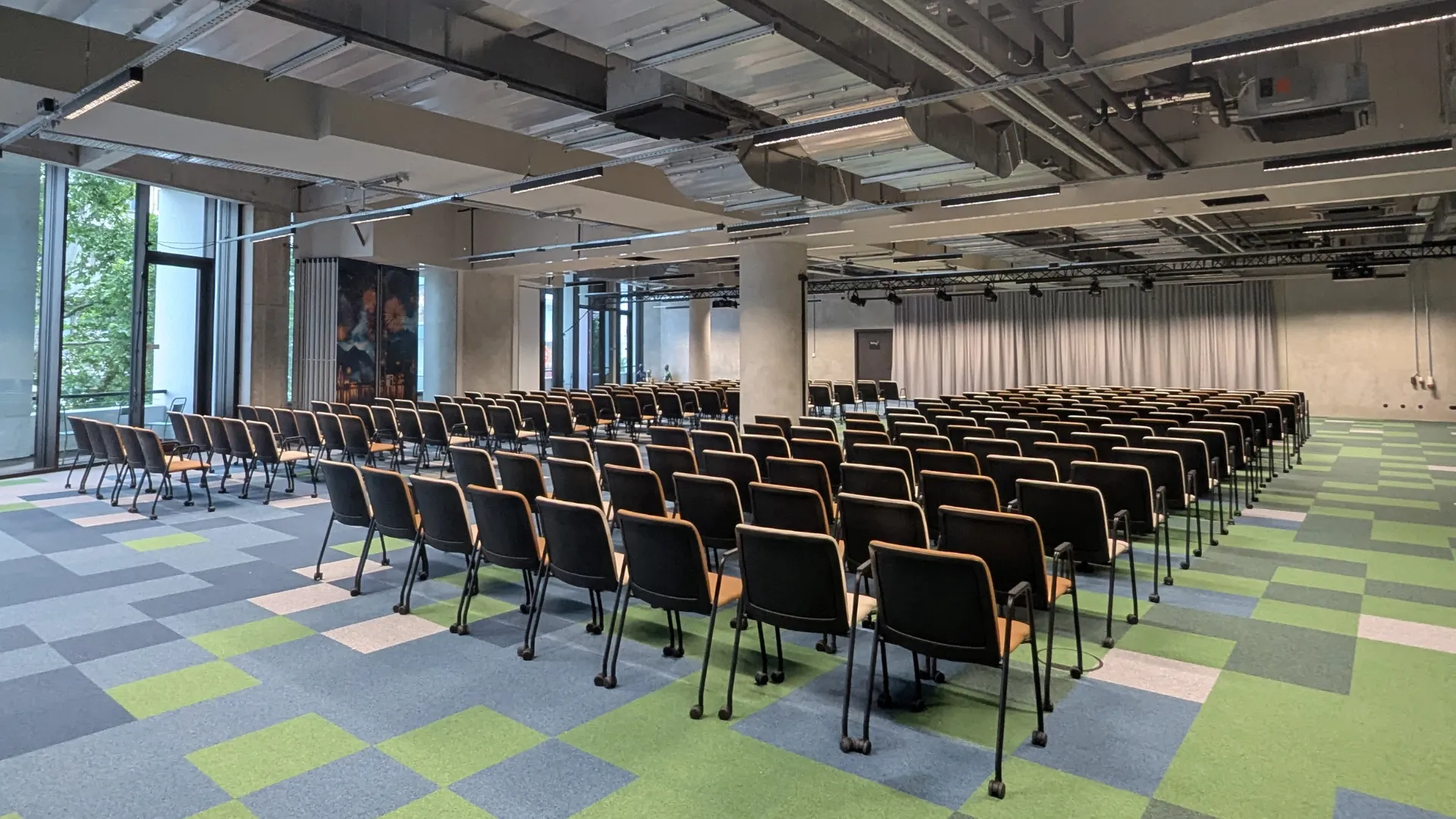 Moderne, große Konferenzhalle mit Reihen von schwarzen Stühlen, grün-blau kariertem Teppich und freiliegenden Deckeninstallationen